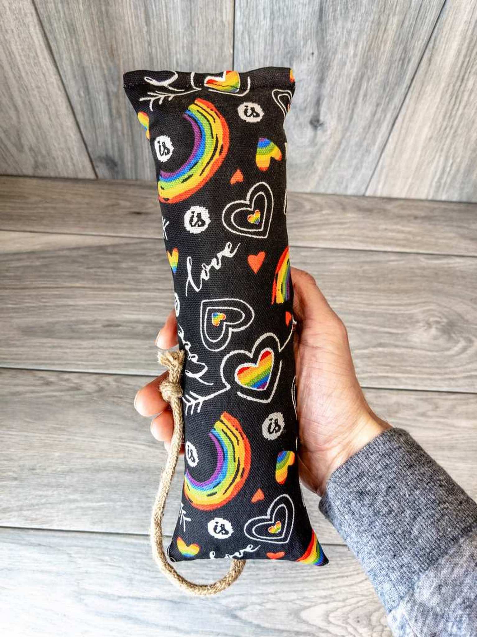 A hand holding a catnip kicker made from a black fabric that has a pattern of rainbows and the word "love" on it.