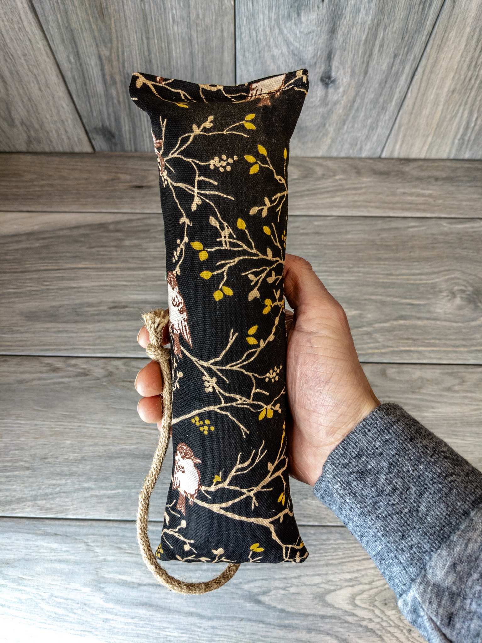 A hand holding a catnip kicker made from a black fabric that has a pattern showing birds sitting in a tree.