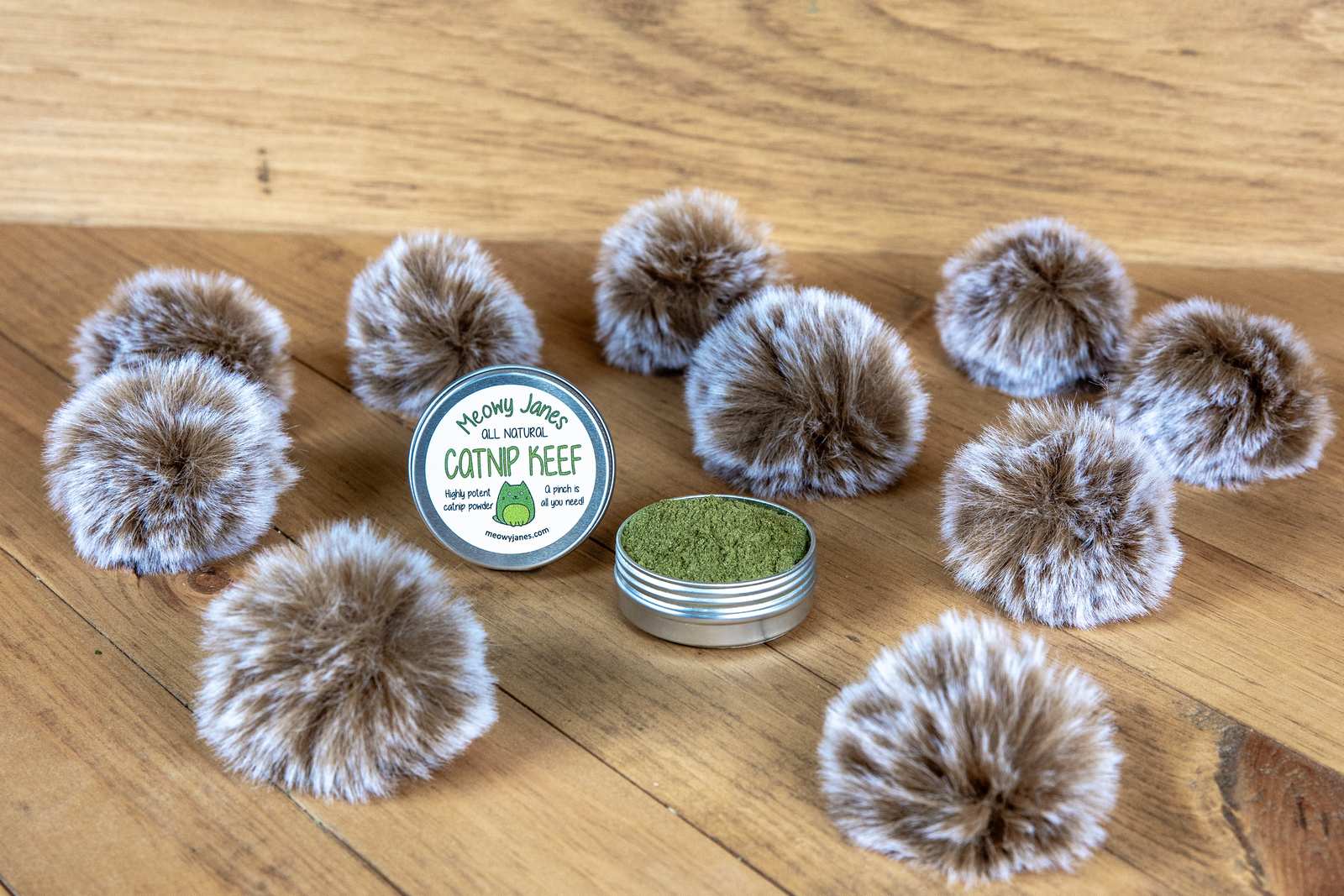 10 grey and brown round cat toys that look like fuzzy pom poms with long hairs. There is a tin of green catnip powder
