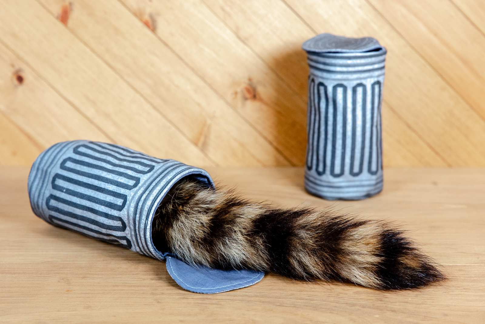 A fabric garbage can cat toy with a raccoon tail sticking out from it.