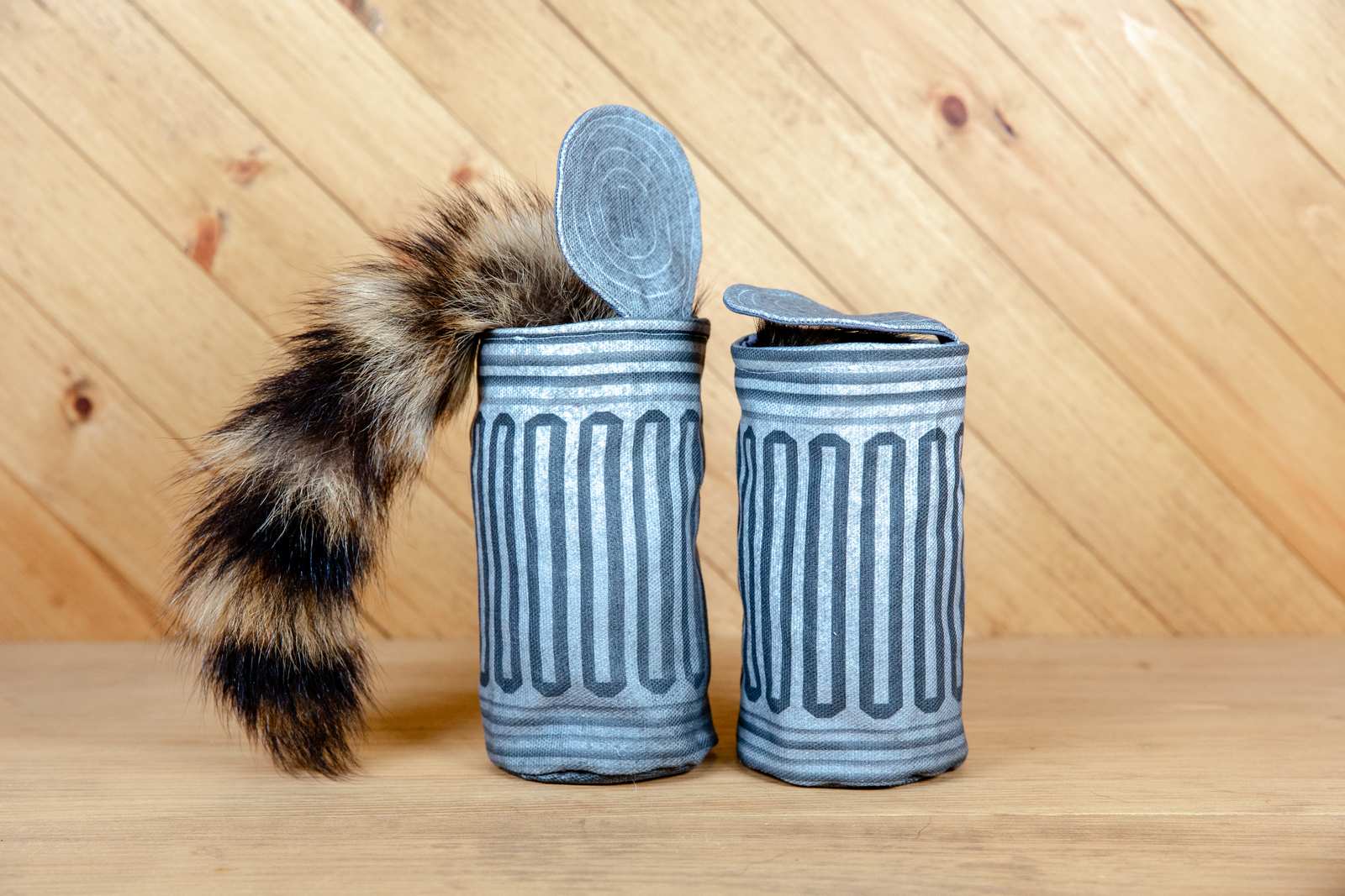 Two fabric garbage can cat toys, one with a raccoon tail sticking out from it and the other with a fabric lid of it.