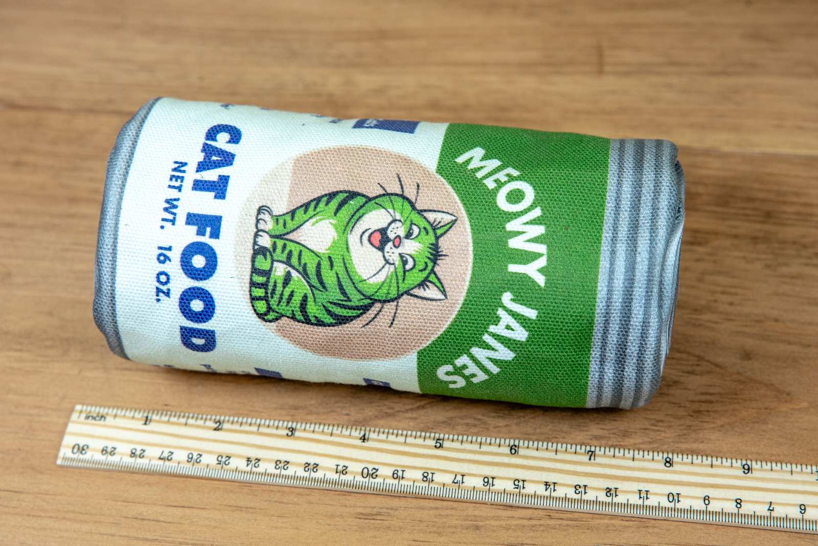 A sewn toy that looks like a can of cat food laying on its side next to a ruler showing it is 8 inches long